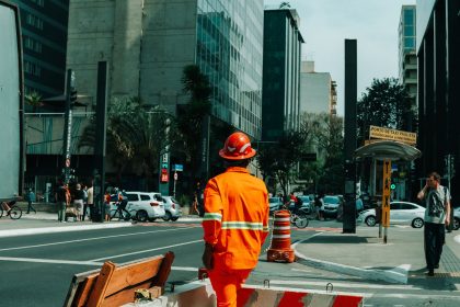 Trabalhador da construção civil em uma rua movimentada de São Paulo, cercado por pedestres e outros elementos da cidade.