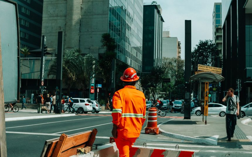 Trabalhador da construção civil em uma rua movimentada de São Paulo, cercado por pedestres e outros elementos da cidade.